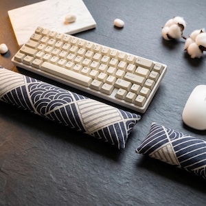 May include: A mechanical keyboard with off-white keys, a white computer mouse, and two fabric wrist rests featuring a dark blue and cream geometric pattern of semicircles and diagonal lines. The items are arranged on a dark grey textured surface.