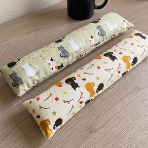 May include: Two rectangular fabric cushions with cat-themed patterns. One cushion features a light green background with white, gray, and calico cat illustrations and paw prints. The other has a cream background with cats and autumn leaves.