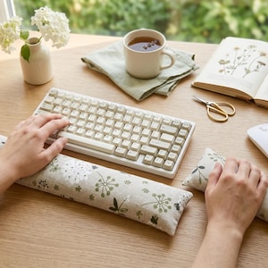 May include: A cream-coloured mechanical keyboard with matching floral-patterned wrist rests. A white mouse and gold scissors are on a wooden desk, with a cup of tea and a book of pressed flowers.