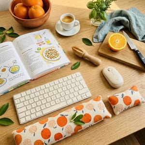 May include: A wooden table setting featuring oranges, a cookbook, and a keyboard. A bowl of oranges, a cup of coffee, and a cutting board with a knife are also present. Orange-themed wrist rests and a computer mouse complete the setup.