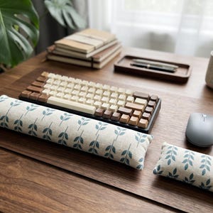 Op de afbeelding: Een houten toetsenbord met crème en bruine toetsen staat op een bureau, naast een bijpassende polssteun en een kleiner kussen, beide met een blauw bladerpatroon. Een grijze muis, een mok en notitieboekjes staan ook op het bureau.