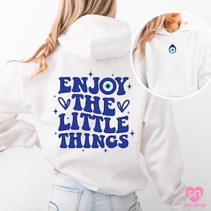 May include: White hoodie with a blue graphic on the back that says "ENJOY THE LITTLE THINGS" with an evil eye symbol. A smaller image of the hoodie shows the evil eye symbol on the front. The hoodie is a casual style.