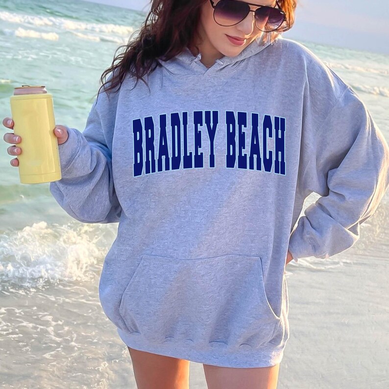 May include: A gray hooded sweatshirt with the text "BRADLEY BEACH" in blue lettering. The sweatshirt is worn by a person standing on a beach.