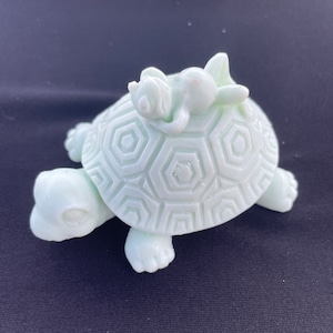 May include: A light green ceramic turtle figurine with a smaller light green ceramic bunny figurine lying on its back on top of the turtle's shell.