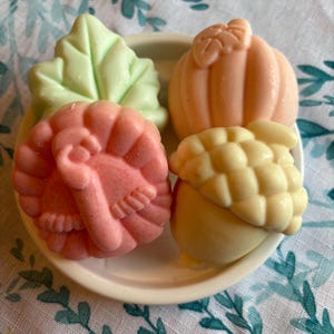 May include: A small, off-white ceramic dish holds four fall-themed soaps. A light green leaf, a peach-colored pumpkin, a pink turkey, and a yellow acorn are arranged on the dish. The soaps are on a white and teal patterned tablecloth.
