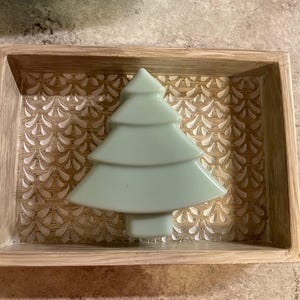 Op de afbeelding: Een lichtgroene keramische kerstboom staat in een houten dienblad met een reliëfpatroon. De bak heeft een lichtbruine afwerking en de boom heeft een gelaagd ontwerp. De achtergrond is een getextureerd oppervlak.