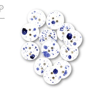 May include: A collection of 12 white round buttons with blue splatter paint. The buttons have two holes each.