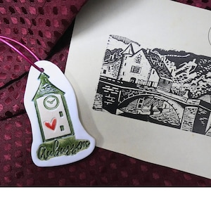 Può includere: Un ornamento a torre dell'orologio in ceramica bianca con tetto verde, un cuore rosso e la parola "Amboise" in verde. Accanto, una stampa in bianco e nero di un ponte e di edifici.