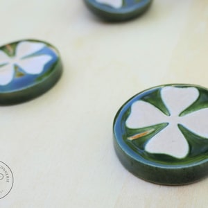 Peut inclure: Trois boutons ronds en céramique avec un vernis vert foncé brillant et un motif de trèfle à quatre feuilles blanc. Chaque bouton a une surface lisse et surélevée. Un logo circulaire avec le texte LA PETITE BOUTONNERIE CERAMIQUES est visible.