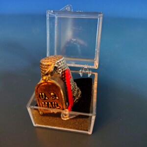 May include: A vintage thimble shaped like a U.S. Mail mailbox, displayed in a clear plastic case. The thimble is bronze-colored with the words "U.S. MAIL" and a red stripe. The thimble is a collectible item.