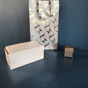 May include: A gift bag with a silver and gold geometric pattern, a plain white box, and a small wooden stamp holder with the word "STAMPS" printed on it. The bag has white rope handles.