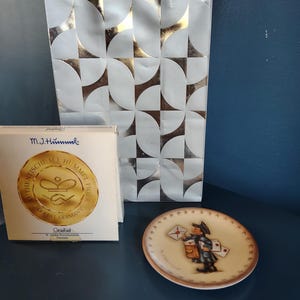 May include: A decorative plate featuring a Hummel figurine of a mail carrier holding letters. The plate has a brown border and a cream-colored background. A box with gold lettering and a silver and white patterned background are also visible.