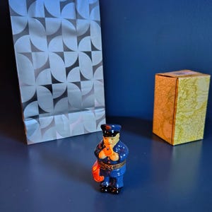 May include: A small, detailed figurine of a mail carrier in a blue uniform, holding a red mailbag. The figurine is on a blue surface, with a patterned gift bag and a small box in the background.