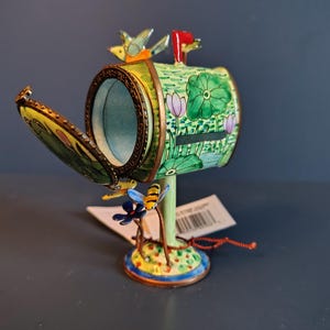 May include: A decorative miniature mailbox with a hinged lid, painted with a garden scene. The mailbox is green with floral and water lily designs, and features a red flag and small bird and bee accents. The base is yellow with a blue and yellow border.