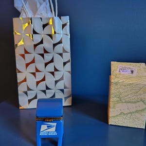 May include: A blue miniature mailbox with the text "UNITED STATES POSTAL SERVICE" is in the foreground. A decorative gift bag with a white and gold geometric pattern and a small box with a map design are also visible.