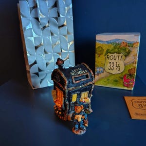 May include: A decorative miniature house with "Local Deliveries" text, a bear figurine, and a Route 33 1/3 box. A silver patterned gift bag is in the background. The items are on a dark blue surface.