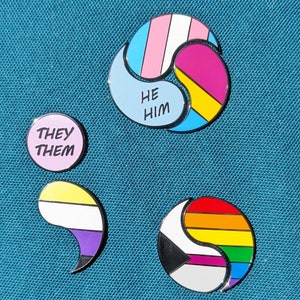 Puede incluir: Cuatro pines de esmalte con diferentes diseños de banderas del orgullo. El pin superior es un símbolo yin-yang con la bandera transgénero en un lado y la bandera bisexual en el otro. El texto "HE HIM" está escrito en el lado azul. El pin izquierdo es un círculo con el texto "THEY THEM" en letras rosas. El pin inferior izquierdo es una bandera genderqueer púrpura, amarilla y blanca en forma de punto y coma. El pin inferior derecho es un símbolo yin-yang con la bandera asexual en un lado y la bandera del arcoíris del orgullo en el otro.
