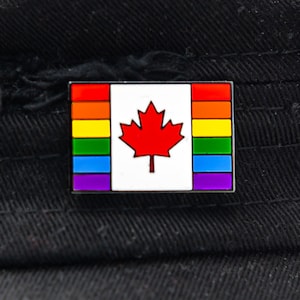 May include: A rainbow-colored Canadian flag pin with a red maple leaf in the center. The pin is on a black fabric background.