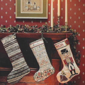 May include: A festive Christmas scene featuring three embroidered stockings hanging from a wooden mantel. Each stocking is personalized with a name: John, Brooke, and Reid. A framed cross-stitch artwork and lit candles complete the holiday decor.