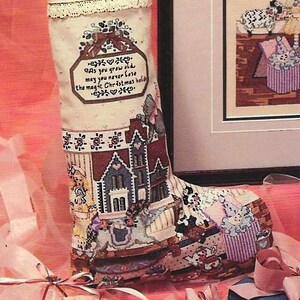 Puede incluir: Un calcetín navideño hecho a mano con el nombre "Adrian" bordado en rojo. El calcetín presenta un diseño de punto de cruz de una escena de pueblo y el texto "As you grow old may you never lose the magic Christmas hold". La parte superior está adornada con encaje y llena de vegetación.