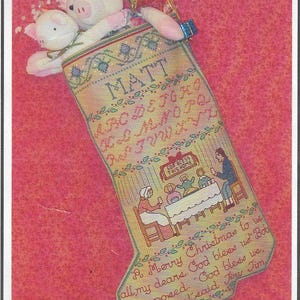 May include: Embroidered Christmas stocking with the name "MATT" and a family scene. The stocking is tan with green and red accents, and features a pig stuffed animal. Text includes "A Merry Christmas to us."