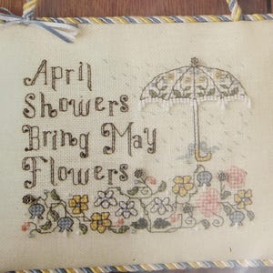 May include: A cross-stitched wall hanging with the text "April Showers Bring May Flowers" and an umbrella design. The embroidery features a floral border with yellow, pink, and blue flowers. The background is a light beige fabric.