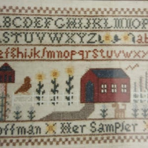 Puede incluir: Sampler de punto de cruz con el alfabeto, una casa roja, un pájaro y flores. El sampler incluye el texto "K.L. Hoffman Her Sampler 1995" y está enmarcado con un borde decorativo.