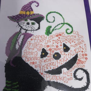 May include: A cross-stitch design featuring a black cat wearing a purple witch hat and a purple and green outfit. The cat is holding a pumpkin with a carved face and Halloween-themed words like "Spook", "Trick or Treat", and "Halloween" in red.
