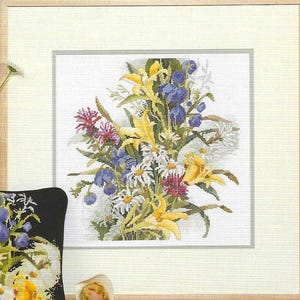 May include: A framed cross-stitch of a floral bouquet. The bouquet features yellow lilies, blue irises, white daisies, and other wildflowers. The artwork is set against a white background and framed in light wood.