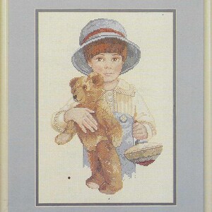 Könnte beinhalten: Kreuzstich-Kunstwerk eines Kindes, das einen Teddybären und einen kleinen Korb hält. Das Kind trägt einen blauen Hut und eine Latzhose. Der Teddybär ist braun. Das Kunstwerk ist in einem hellgrauen Rahmen gerahmt.