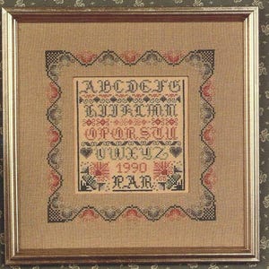 May include: Framed cross-stitch sampler with a tan background and a gold-colored frame. The sampler features the alphabet, numbers, and the year 1990. The design includes red, black, and blue decorative elements.