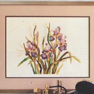 May include: A framed cross-stitch of irises in shades of purple, green, and yellow. The artwork is set against a cream-colored background and framed with a light brown wooden frame and a pink matting.