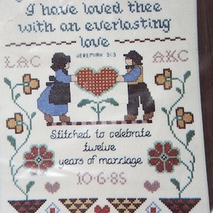 May include: Cross-stitch artwork celebrating twelve years of marriage, featuring two figures holding a heart. The text reads "Yea, I have loved thee with an everlasting love" and "Stitched to celebrate twelve years of marriage 10-6-85."