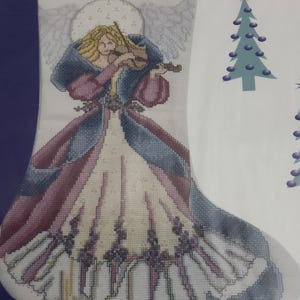 May include: Cross-stitch Christmas stocking featuring an angel playing a violin. The angel has blonde hair, a halo, and wears a long gown in shades of white, pink, and blue. The name "KARY" is stitched at the top.