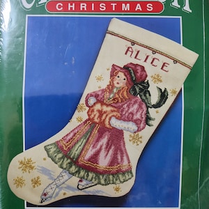 May include: Cross stitch Christmas stocking kit featuring a Victorian skater design. The stocking is cream-colored with a detailed image of a girl in a pink dress and ice skates. The name "ALICE" is stitched at the top. The kit measures 9" x 16".