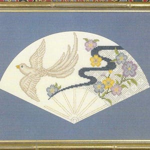 May include: Cross-stitch artwork of a fan design. The fan features a flying bird, a black ribbon, and floral accents in yellow, blue, and purple. The fan is set against a blue background and framed in gold.