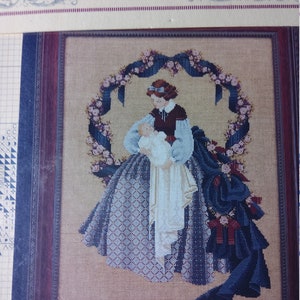 Op de afbeelding: Kruissteekpatroon met een vrouw in een lange jurk die een baby vasthoudt, omringd door een bloemenkrans. Het kunstwerk is omlijst in een donkere houten lijst, met de tekst "Lavender & Lace" bovenaan.