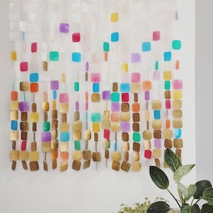 Op de afbeelding: Een kleurrijke wanddecoratie gemaakt van iriserende en gouden vierkanten. De vierkanten zijn in een cascade-patroon aan elkaar geregen, wat een modern en abstract ontwerp creëert.