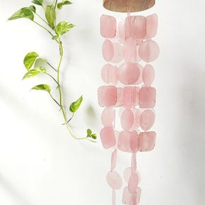 May include: A pink wind chime with a wooden top and a series of pink, square and round, translucent shapes hanging below. A green leafy plant is to the left of the wind chime against a white background.