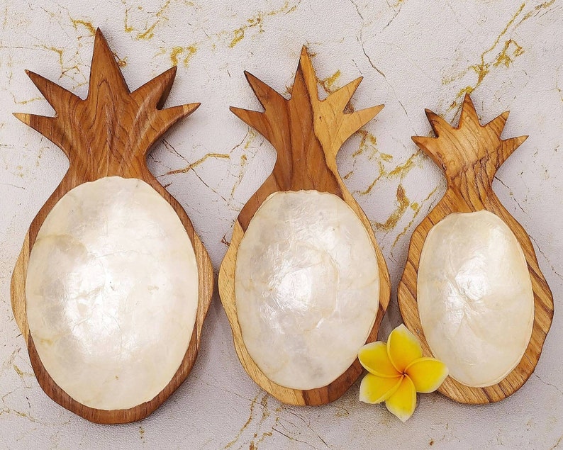 May include: Three wooden pineapple shaped bowls with white mother of pearl interiors. The bowls are different sizes and are arranged in a row on a light brown surface.