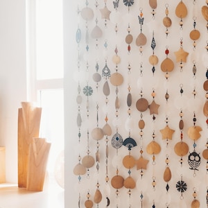 May include: A decorative hanging room divider composed of multiple vertical strands featuring a variety of shapes including stars, circles, teardrops, and intricate metal charms in gold, white, and black tones, interspersed with small beads.