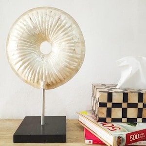 May include: A white, round, decorative object with a hole in the centre, mounted on a black stand. The object is made of a material that resembles mother of pearl.