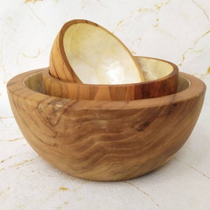 May include: A set of three nesting bowls. The largest bowl is made of natural wood with a brown and tan grain pattern. The two smaller bowls are made of white mother of pearl with a subtle iridescent sheen.
