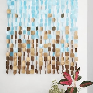 Op de afbeelding: Een wanddecoratie gemaakt van blauwe en gouden vierkante stukjes parelmoer. De stukjes zijn aan een houten stok bevestigd en hangen in een watervalpatroon.