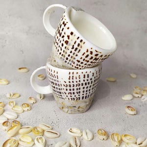 Puede incluir: Una taza de cerámica blanca con un asa negra y el texto "But First, Coffee" en un estilo manuscrito. La taza está sobre una superficie blanca. La taza mide aproximadamente 10 cm de alto.