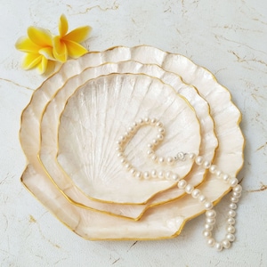 Puede incluir: Tres bandejas blancas en forma de concha con ribetes dorados, apiladas una encima de la otra. Un collar de perlas está drapeado sobre la bandeja superior.