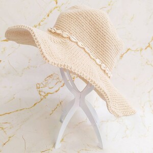 May include: A cream-colored, wide-brimmed crochet hat adorned with small seashells along the brim. The hat is displayed on a white stand against a marble-patterned background. The hat's brim is wavy, and the crochet work is visible.