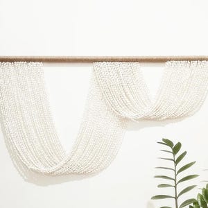 以下が含まれることがあります： 木製の棒に白いビーズが飾られた壁掛け。ビーズは滝のように連なっており、モダンでミニマルな印象を与えます。