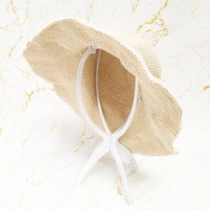 May include: A beige woven sun hat with a wide, wavy brim. The hat is displayed on a white hat stand. The hat has a textured appearance and is designed to provide shade from the sun.