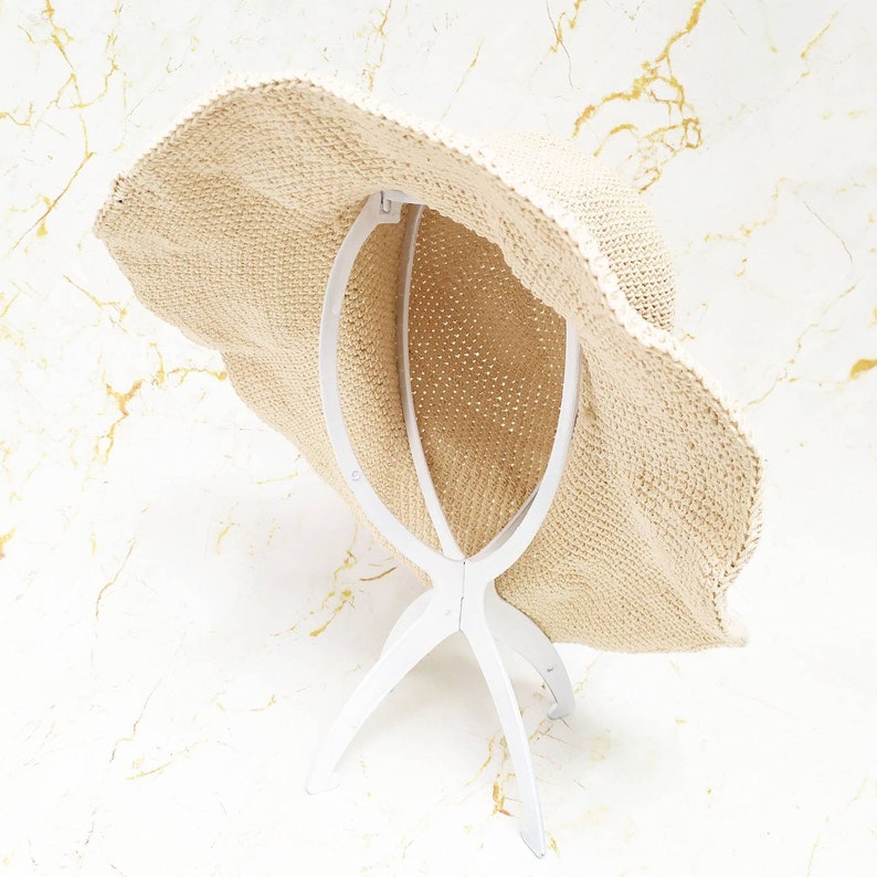 May include: A beige woven sun hat with a wide, wavy brim. The hat is displayed on a white hat stand. The hat has a textured appearance and is designed to provide shade from the sun.